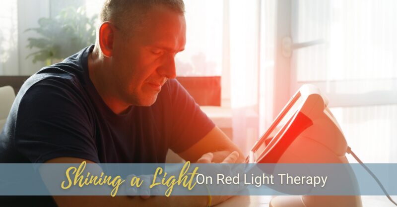 An older man sits at a table with his eyes closed as a red light therapy panel device beams red light onto his face.