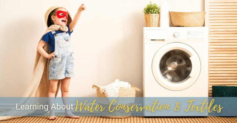 A young boy wearing a red face mask and a towel as a cape is pointing up and away next to a washing machine.